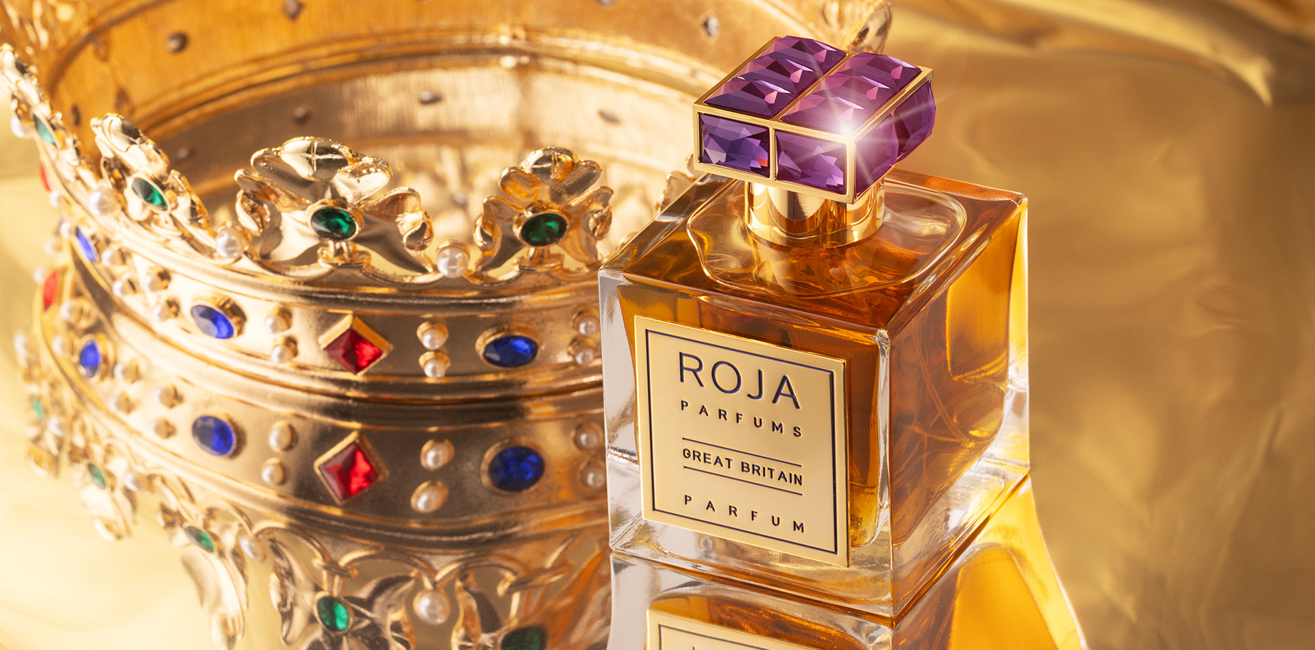 A bottle of Roja Parfums Great Britain perfume sits on a reflective gold surface beside an ornate golden crown decorated with colorful jewels. The scene exudes luxury and elegance.