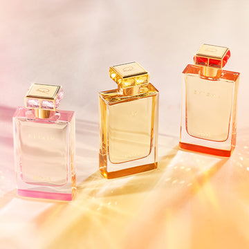 Three elegant perfume bottles labeled ELIXIR and ROJA, displayed on a light surface with warm, pastel lighting creating soft reflections and a luxurious atmosphere.