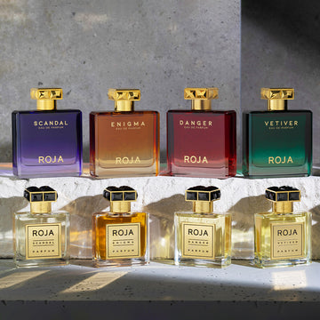 Six Roja perfume bottles are displayed: four square bottles labeled Scandal, Enigma, Danger, and Vetiver on a stone ledge, with their matching smaller gold bottles lined up in front of them.