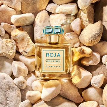 A bottle of Roja Isola Blu Parfum with a blue gem-like cap is placed on a bed of smooth, light-colored rocks. The label on the bottle is blue and gold.