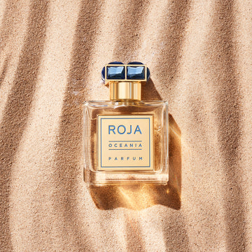 A bottle of Roja Oceania Parfum with a gold cap and label lies on sunlit sand, casting a soft shadow.