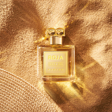 A gold bottle of Roja Isola Sol Parfum rests on textured golden sand, with sunlight creating shiny reflections around it. Part of a woven sun hat appears in the lower left corner.