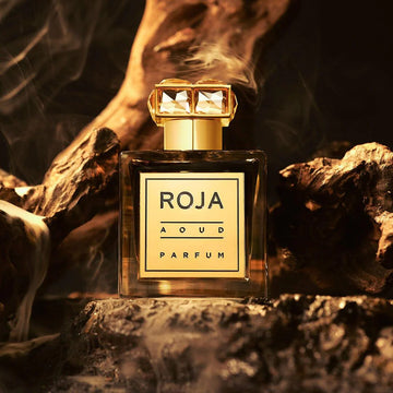 A bottle of Roja Aoud Parfum with a gold cap is displayed between twisted, dark wood and surrounded by smoky, warm lighting, creating a luxurious and exotic atmosphere.