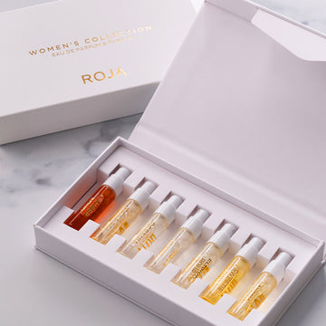 A white box labeled The Womens Discovery Collection by Roja Parfums displays seven luxurious fragrance vials in yellow and amber hues, neatly arranged in a white tray on a marble surface.