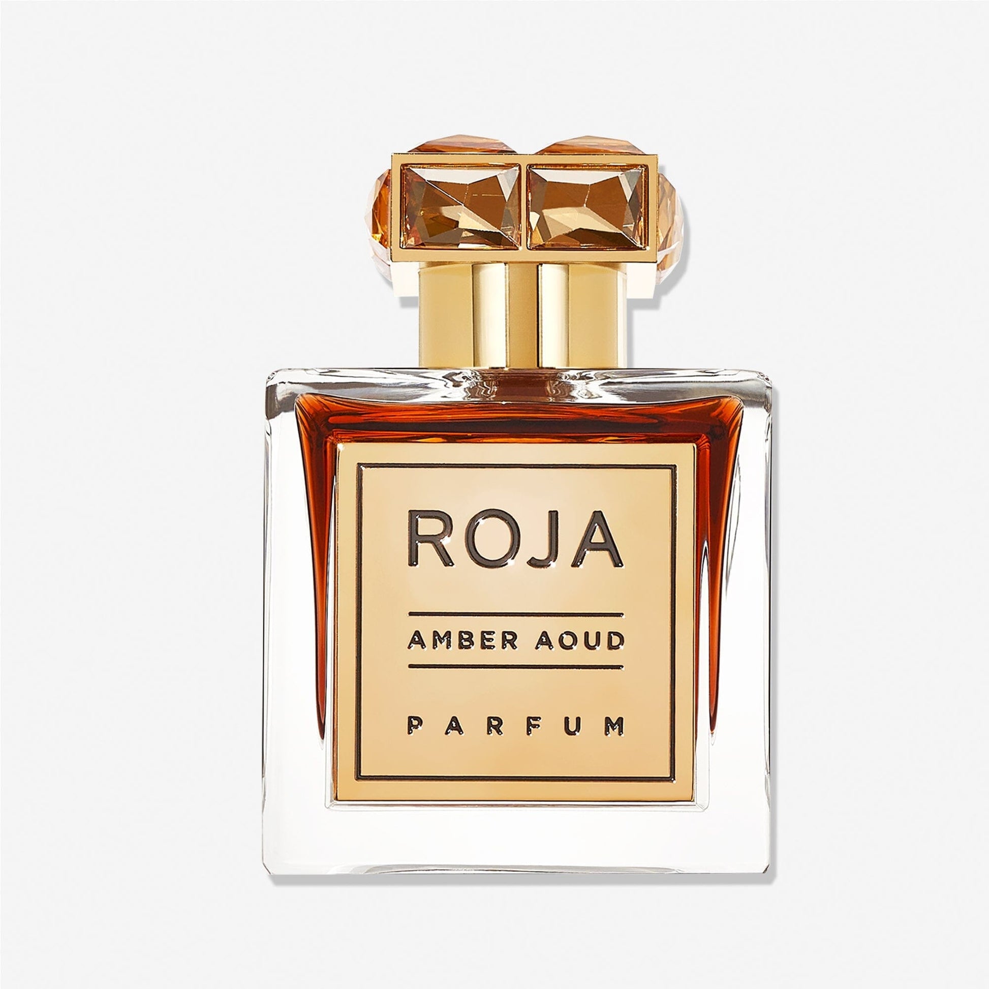 A clear perfume bottle with an ornate gold-capped stopper holds amber liquid. The gold label reads Amber Aoud by Roja Parfums, highlighting oud-enriched notes.