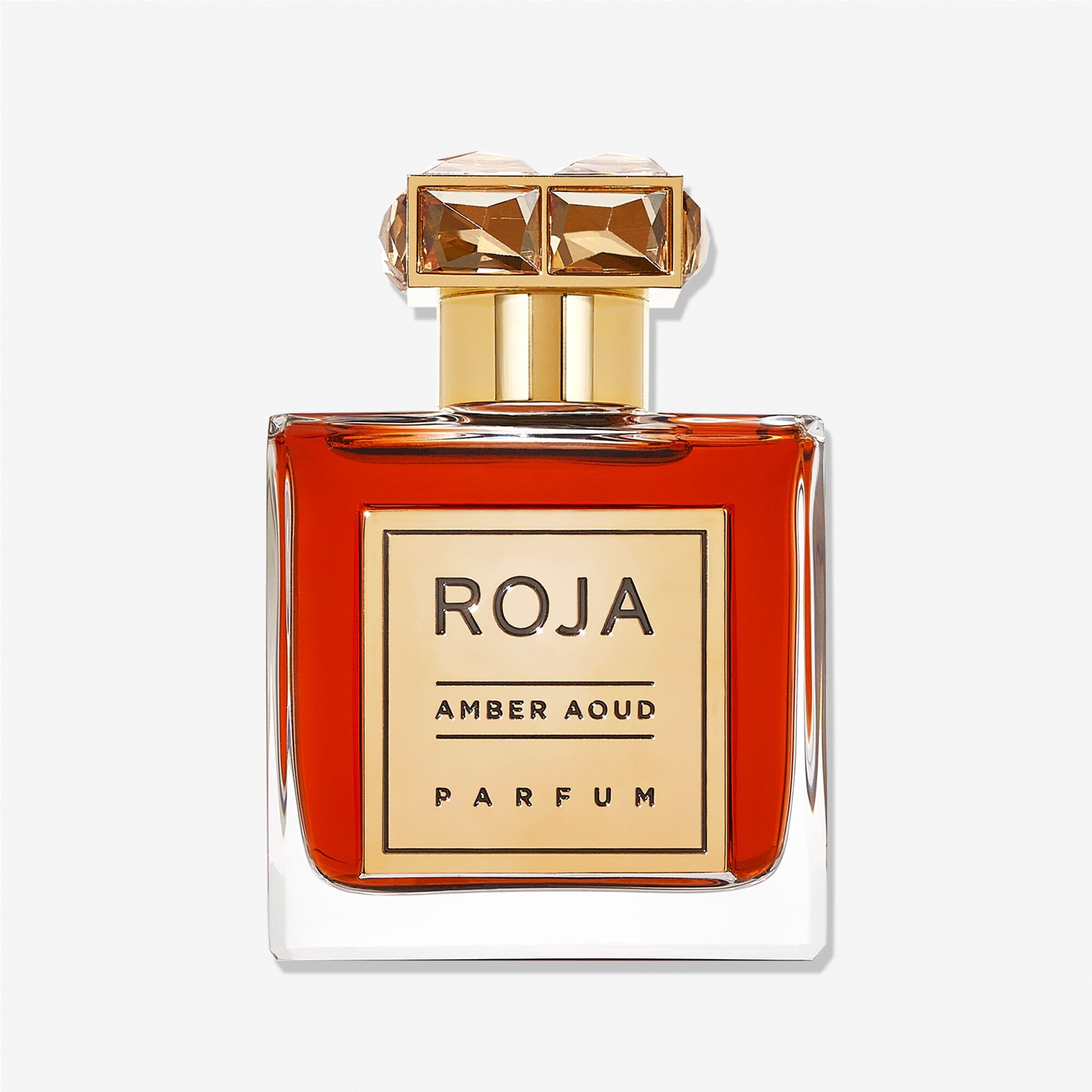 A clear glass bottle of Roja Parfums Amber Aoud, featuring a gold cap and label and filled with deep amber liquid, is set against a white background—showcasing the luxury of oud in every drop.