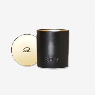 A black Amber Aoud candle holder by Roja Parfums, embossed with ROJA on the front, sits beside its matching gold lid featuring a decorative “R” emblem—an elegant display reminiscent of olfactive majesty, set against a plain white background.