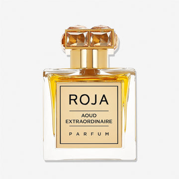 A square glass bottle of Roja Parfums Aoud Extraordinaire holds golden-yellow liquid, featuring a gold gem-accented cap and a front label reading AOUD EXTRAORDINAIRE, capturing the essence of refined oud.