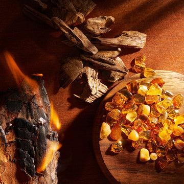 A close-up of polished amber stones on a round wooden plate, dried wood pieces above it, and a burning log with visible flames on the left, all set on a brown surface.