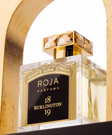 A clear glass bottle of Roja Parfums 18 Burlington 19 perfume with a decorative gold and black label and a gold, gem-like cap, displayed on a gold shelf with a white background.