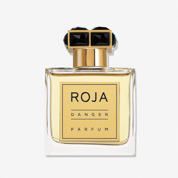 A clear glass bottle filled with yellow liquid, labeled Danger Pour Homme by Roja Parfums in bold black on gold, features a gold cap with two black gem-like shapes and embodies a seductive mens fragrance with an irresistible scent.