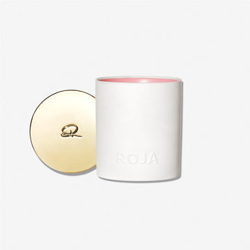 A white Roja Parfums candle, “Dawn In The Rose Garden,” features pink wax and ROJA embossed on the front. Its gold lid, adorned with a stylized R, rests beside it against a simple, light background.
