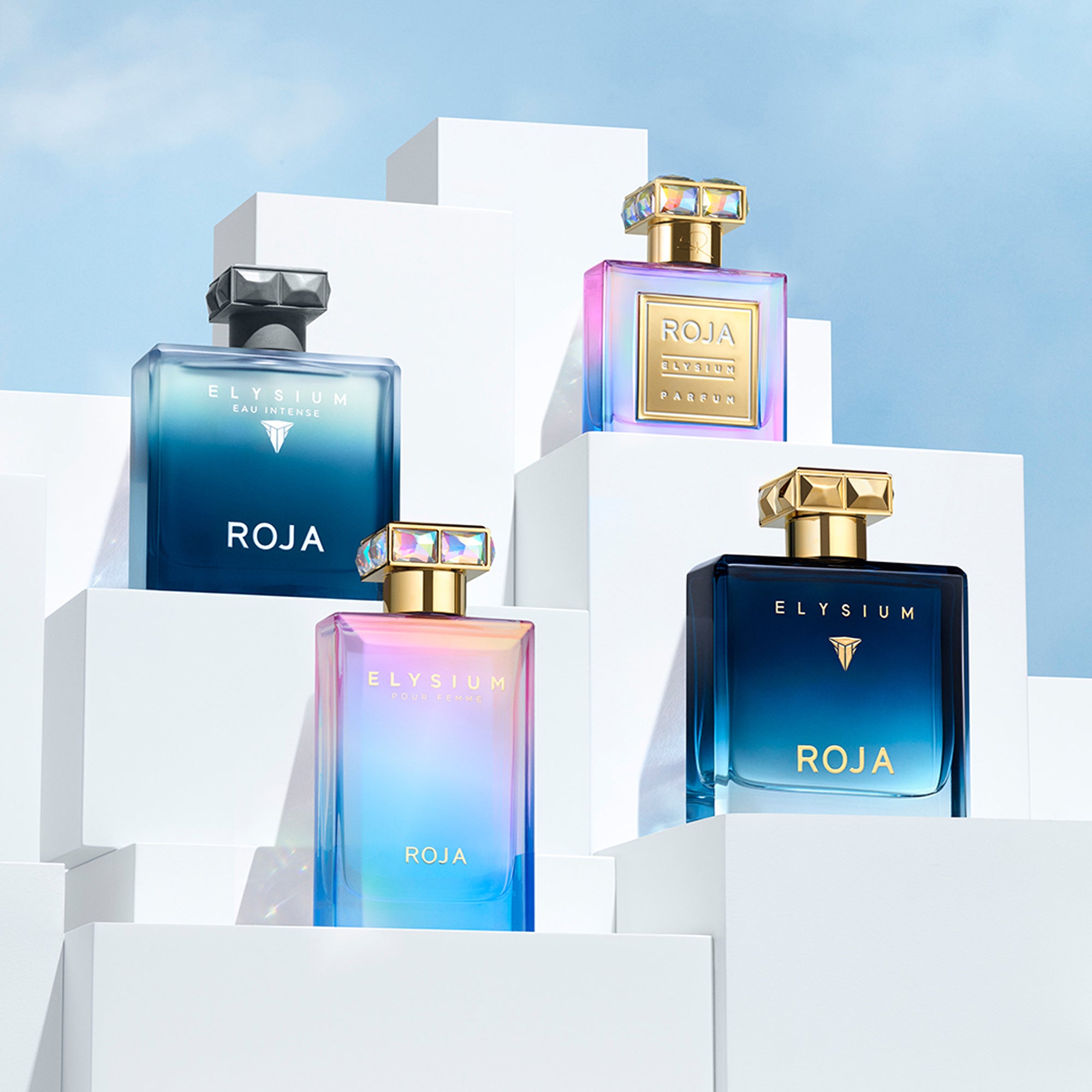 Three bottles of Roja Elysium perfume, each with a geometric cap, are displayed on white, stepped blocks against a blue sky. The bottles have blue and iridescent gradients with gold and silver accents.