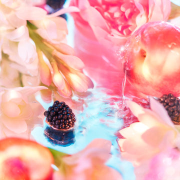 The Elysium Pour Femme Coffret by Roja Parfums captures an ethereal scene with a close-up of peach, blackberries, and pastel flowers amid glistening water. Its refreshing blend of pink, white, and light blue tones crafts a delicate and vibrant aesthetic.
