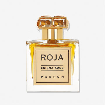 A clear glass perfume bottle of Enigma Aoud by Roja Parfums, featuring golden accents and a crystal-like gold cap, filled with amber-hued scent, is displayed against a plain white background.