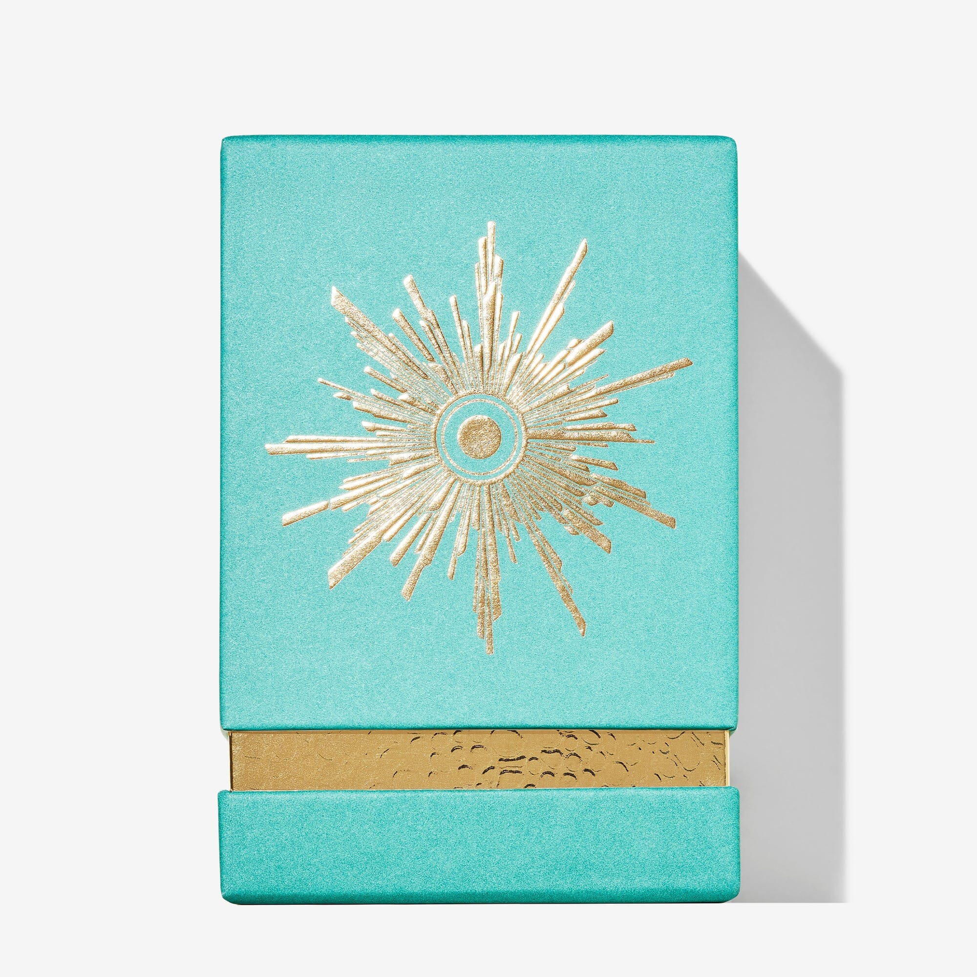 A turquoise rectangular Fortnum & Mason box by Roja Parfums features a gold, embossed sunburst on the lid and a textured gold accent along the bottom edge, inspired by an Ambrée perfume. It sits on a white background.