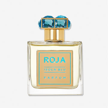 A clear glass bottle of Roja Parfums Isola Blu features a gold label with turquoise text, topped by a gold cap accented with two large blue gemstones, evoking the luxury of a five-star getaway.