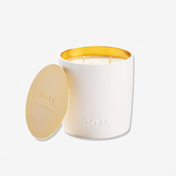 A white candle named London by Roja Parfums sits in a white container with a gold interior. Its matching gold lid, embossed with ROJA PARFUMS, rests beside it—offering an elegant tribute to Londons timeless hedonism.