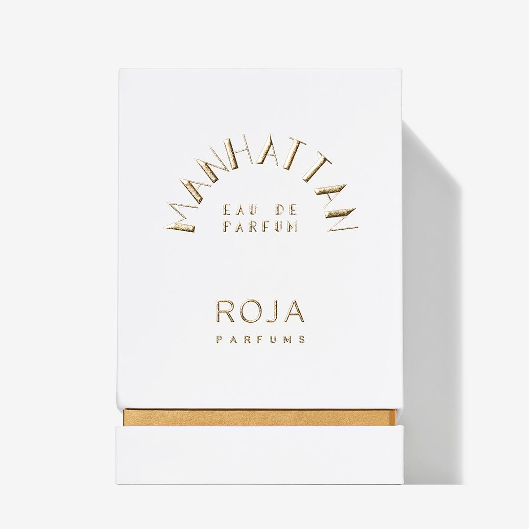 A white box with gold text displays the Manhattan fragrance by Roja Parfums, featuring a gold accent at the base. The sleek, elegant packaging mirrors the sophisticated urban essence of the scent inside.