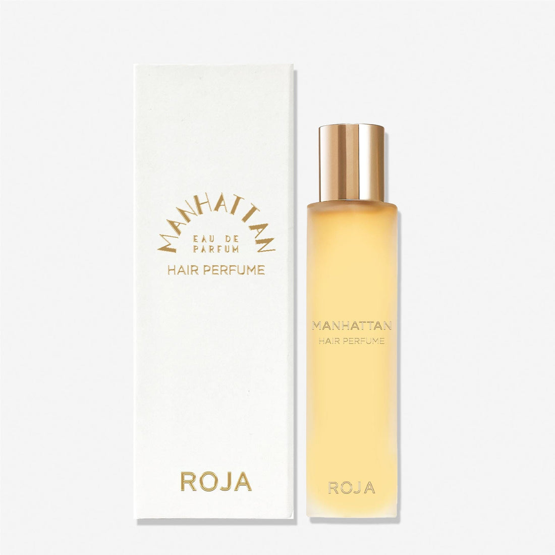 A gold-capped bottle of Roja Parfums Manhattan Hair Mist stands next to its white box, both with gold text and a minimalist design on a light background, evoking the luxurious scent experience of cedarwood.