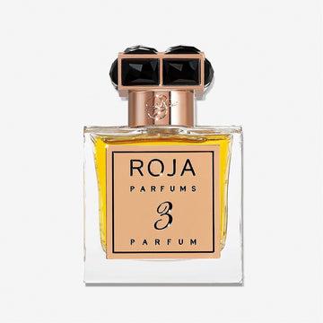 A clear glass bottle with golden-yellow liquid labeled Roja Parfums Parfum De La Nuit 3 features a square gold cap topped with two large black gemstones, evoking the exotic allure of the Parfum De La Nuit collection.