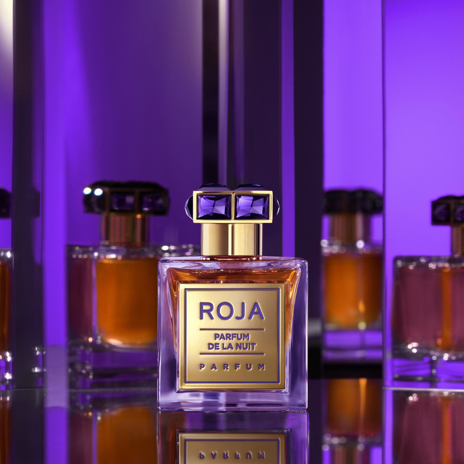 A bottle of Parfum De La Nuit by Roja Parfums rests on a reflective surface, surrounded by mirrors and bathed in purple light, creating striking reflections of this sensual fragrance.