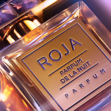 Close-up of a luxurious perfume bottle with a gold label reading ROJA PARFUM DE LA NUIT PARFUM. The bottle has a faceted cap and a warm, glowing light reflecting off the glass.