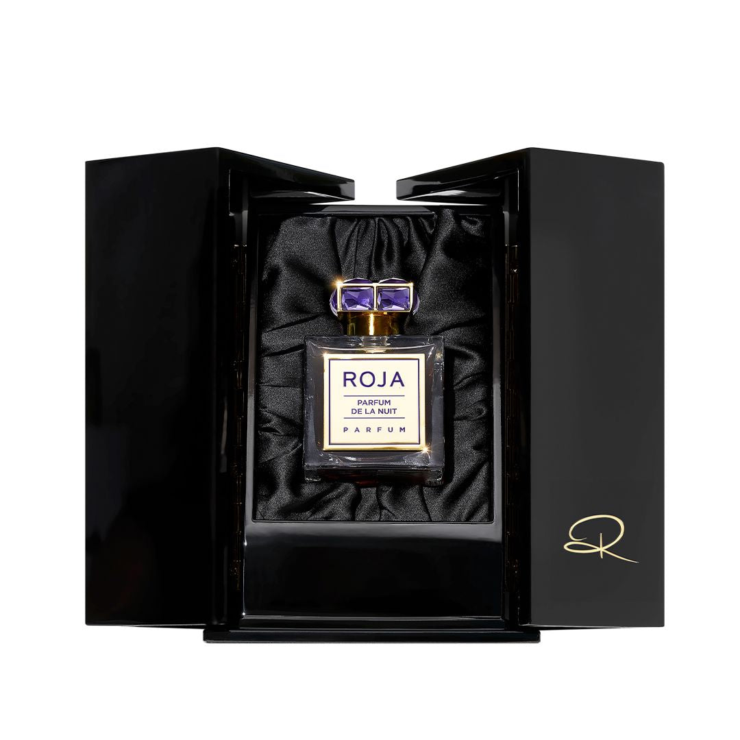 A bottle of Parfum De La Nuit by Roja Parfums sits in an open glossy black box with black fabric lining, gold label, and a faceted purple and gold cap. The box features a gold signature.