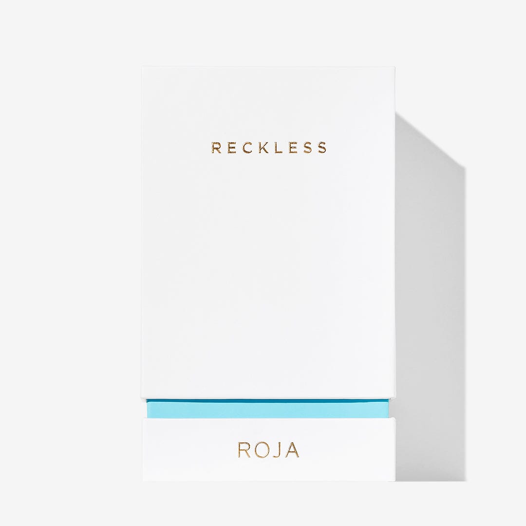 A white rectangular box displays RECKLESS in gold at the top and ROJA in gold on a blue band at the bottom, representing Roja Parfums Reckless Pour Femme, evoking a summery scent with Neroli and Sandalwood notes on a plain white background.