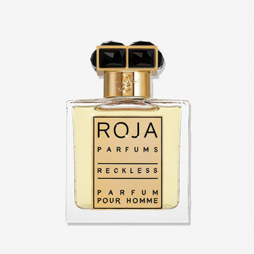 A clear glass bottle of Roja Parfums Reckless Pour Homme features a gold and black cap and holds light yellow liquid. The label shows the brand and product name in bold black letters—a masculine scent with spicy chypré notes.