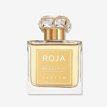 A clear glass bottle filled with golden liquid, labeled Scandal Pour Femme by Roja Parfums. The gold cap with two large square gemstones hints at a bold blend of white flowers and sandalwood musk. Set against a plain white background.