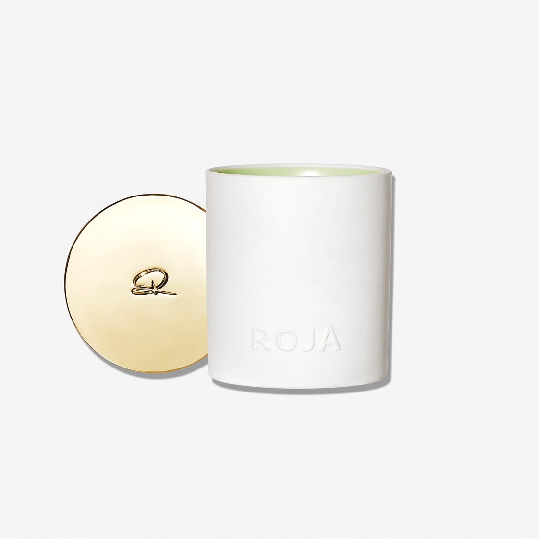 A white Strolling Through The Orchard candle by Roja Parfums, ROJA embossed on the front, sits with its round gold lid beside it. Inspired by orchard charm and crisp fruit scents, the elegant R logo accents the lid on a plain white background.
