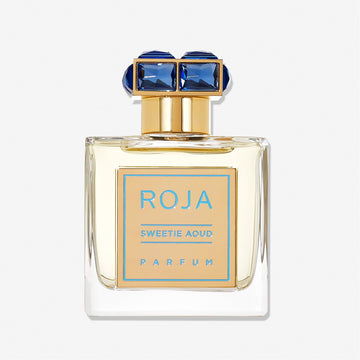 A clear bottle of Roja Parfums Sweetie Aoud, featuring gold accents and a blue gemstone-studded cap, captures the essence of a gourmand and sweet oud perfume, showcased against a pristine white background.