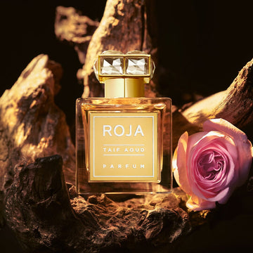 A square glass bottle of Roja Parfums Taif Aoud with a faceted gold cap sits on textured wood. Its label reads TAIF AOUD. A pink rose lies beside it, while warm light, echoing frankincenses glow, completes the scene.