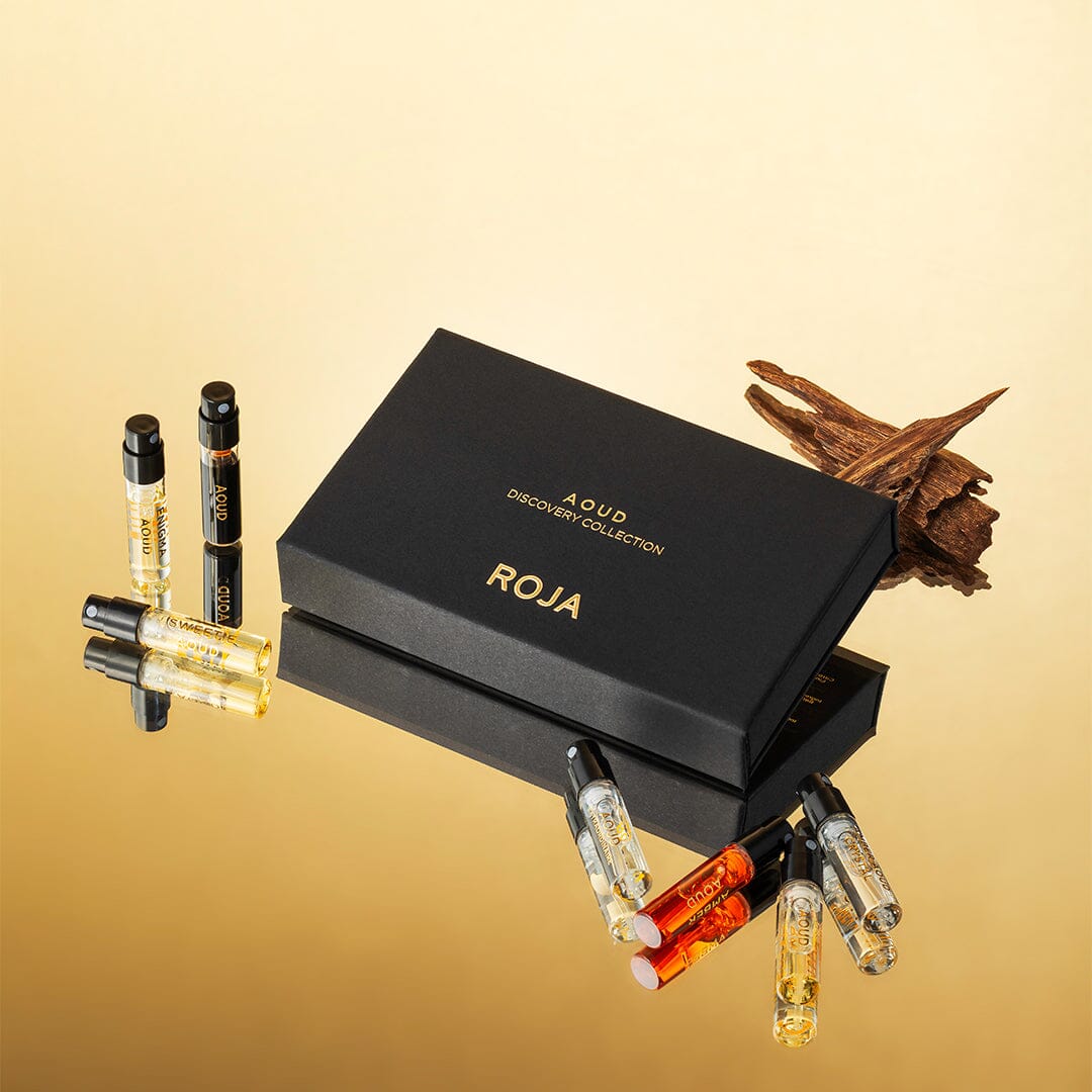 A black box labeled The Aoud Discovery Collection by Roja Parfums, inspired by Arabian scents, is surrounded by sample perfume vials and oud wood, all displayed against a golden backdrop.