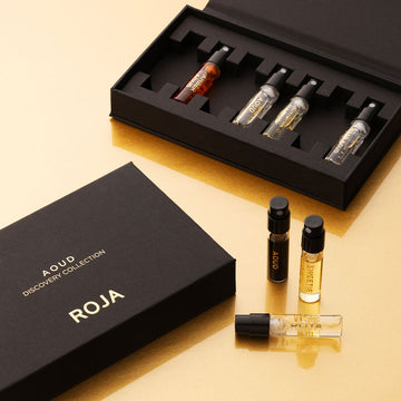 A black box labeled The Aoud Discovery Collection by Roja Parfums contains five perfume vials with luxurious oud scents. Three vials are shown on a gold surface, accented by elegant gold and white text.