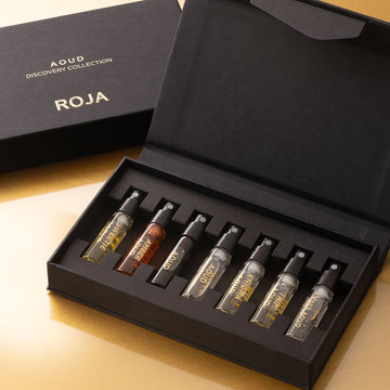 The Aoud Discovery Collection by Roja Parfums features seven glass perfume spray vials, each labeled with a unique scent—like Amber Aoud—elegantly presented on gold and showcasing the rich Oud ingredient.