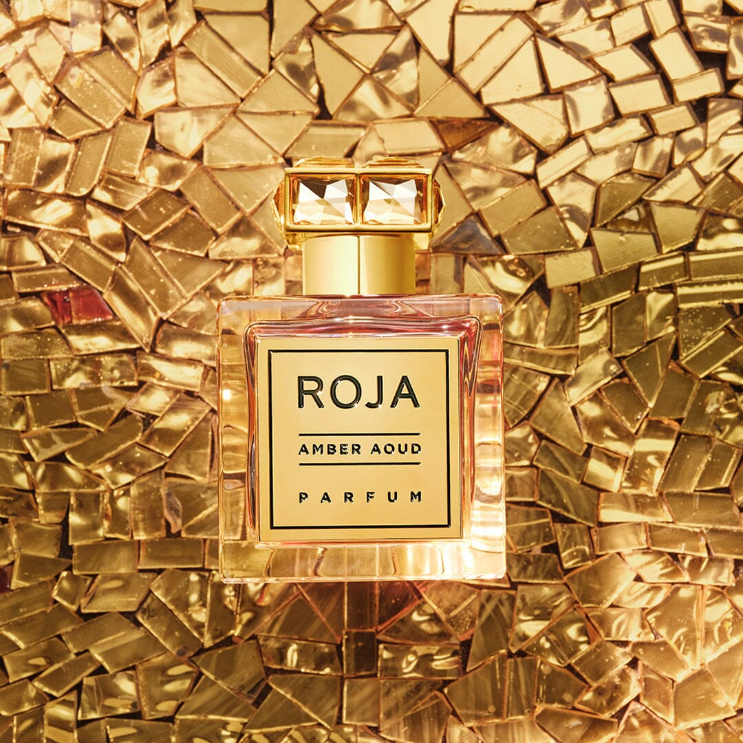 A bottle of Roja Amber Aoud Parfum sits on a mosaic of shiny gold tiles, with golden light reflecting off both the bottle and the background.