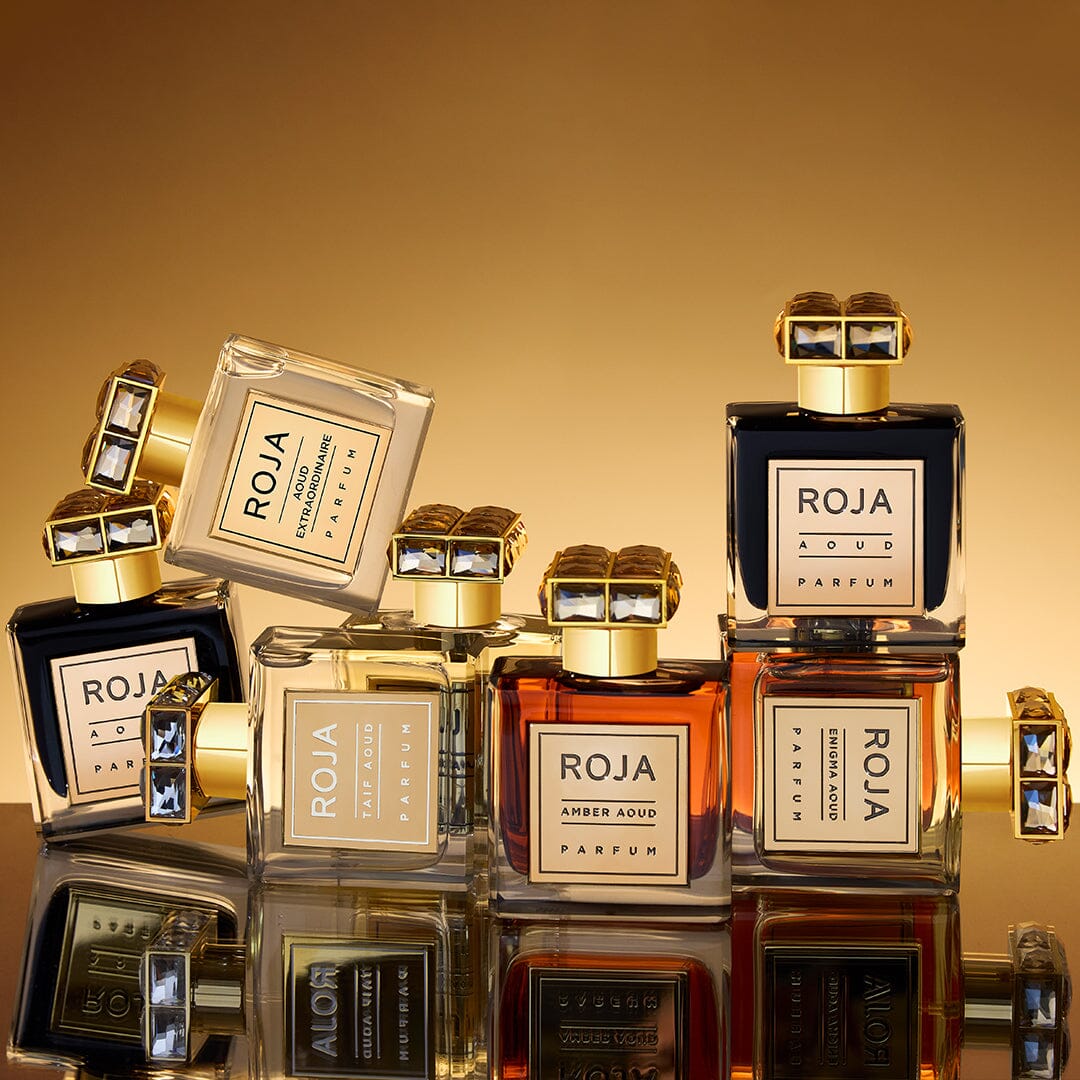 A group of luxurious Roja Parfums bottles in various colors, stacked and arranged against a shimmering gold background, with elegant gold caps and reflective surfaces enhancing the upscale presentation.