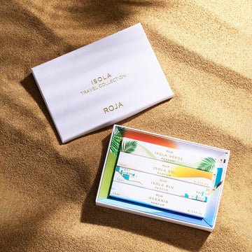 An open box of The Isola Travel Collection by Roja Parfums rests on sand, revealing five vibrant fragrance vials in a tray adorned with tropical designs—an invitation to escape with the Isola Collection.