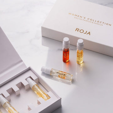 An open white Roja Parfums box labeled The Womens Discovery Collection reveals sample vials. Three vials stand upright nearby, each containing exquisite Roja Parfums fragrances in various colored liquids.