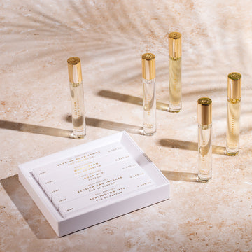 Five glass perfume vials with gold caps stand on a beige surface next to an open white box holding five more labeled perfume vials. Soft shadows and sunlight create a warm, elegant atmosphere.
