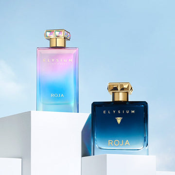 Two Elysium Roja perfume bottles sit on white platforms against a blue sky—one bottle is iridescent pastel colors with a jewel-like cap, the other is dark blue with a gold cap.
