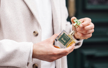 A person in a light-colored coat holds a bottle of Roja Parfums perfume, unscrewing the green and gold cap. The background is slightly blurred, focusing on the hands and fragrance bottle.