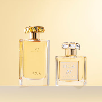 Two luxury perfume bottles labeled ROJA 51 stand on a beige surface against a soft gold background; one bottle is tall and rectangular, the other is shorter and square. Both have gold-toned caps accented with crystals.