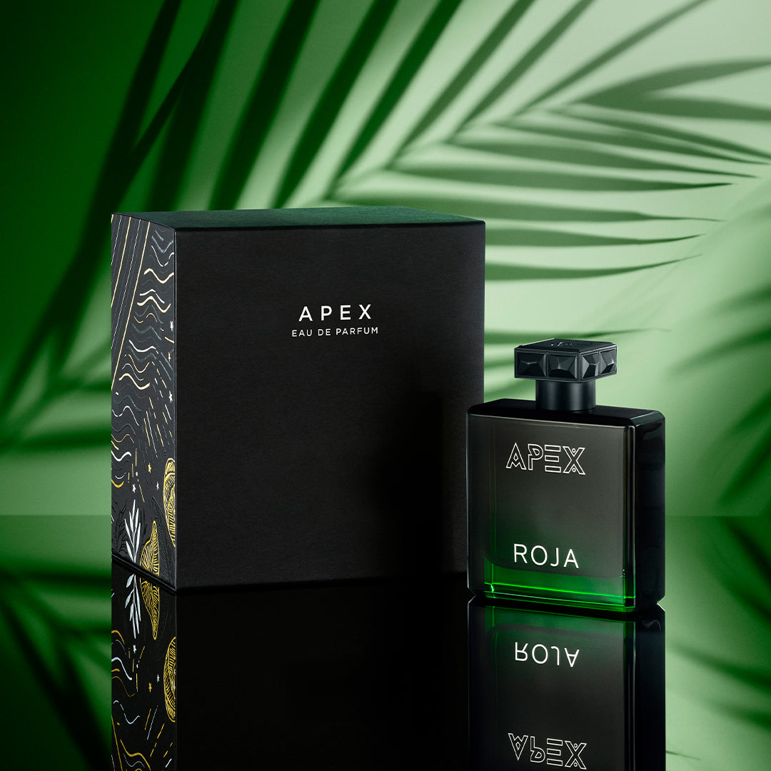 A black bottle of Roja Apex Eau de Parfum stands next to its matching black box, both set against a green background with palm leaf shadows. The bottle features a green gradient at the base and bold white text.