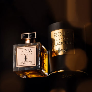 A bottle of Roja Parfums The Exclusive Black Tier perfume stands upright next to its matching black and gold cylindrical case, both with elegant gold labels, against a dark, luxurious background.