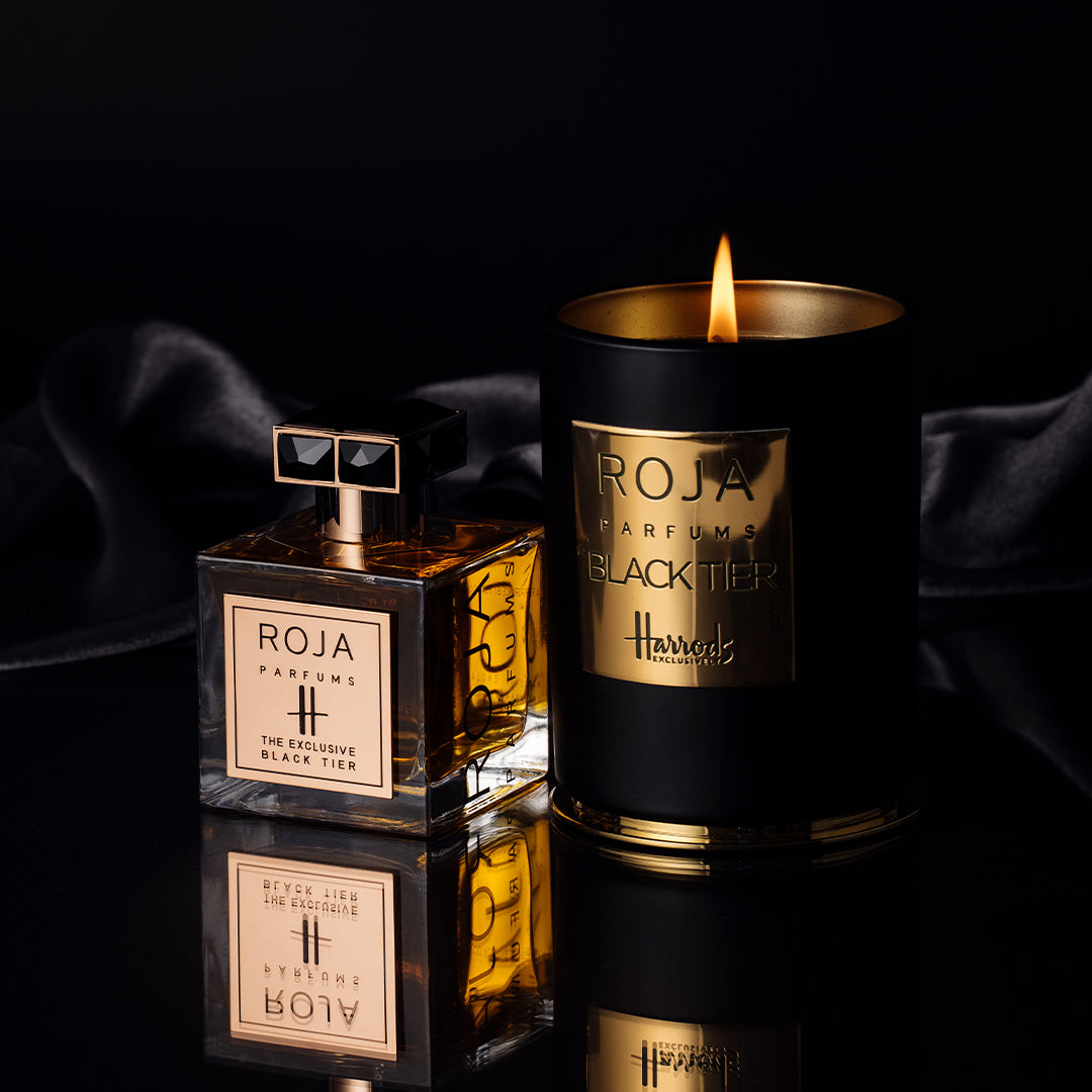 A luxurious scene featuring a bottle of Roja Parfums The Exclusive Black Tier perfume beside a lit black and gold Roja Parfums candle, set against a dark, reflective surface with a draped black fabric in the background.