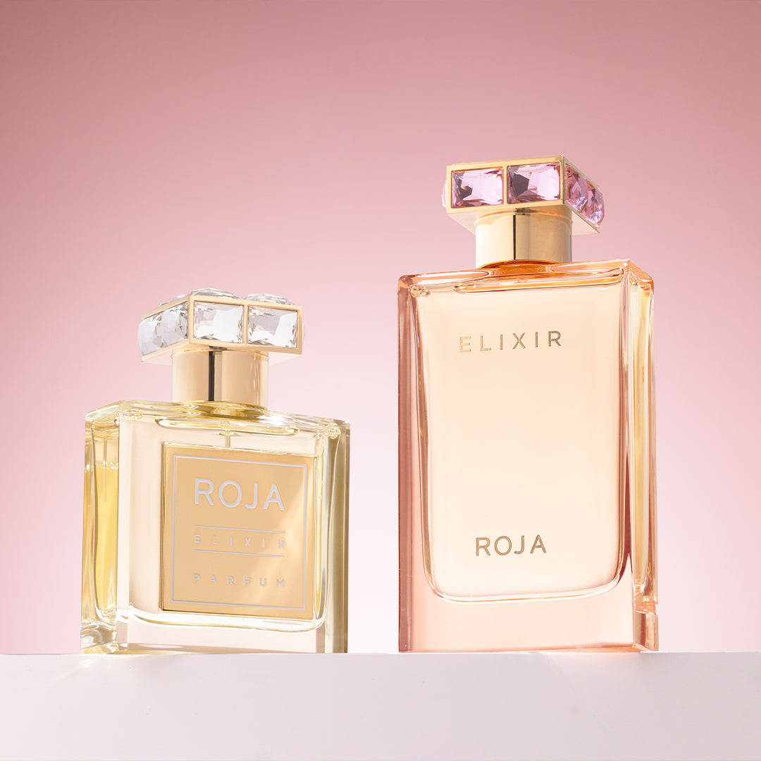 Two elegant perfume bottles labeled ROJA ELIXIR are displayed against a soft pink gradient background. The bottles have square jeweled caps, with the left bottle smaller and filled with golden liquid.