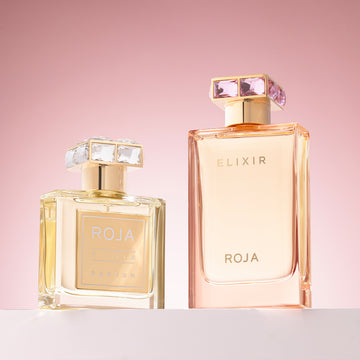 Two elegant perfume bottles labeled ROJA ELIXIR are displayed against a soft pink gradient background. The bottles have square jeweled caps, with the left bottle smaller and filled with golden liquid.